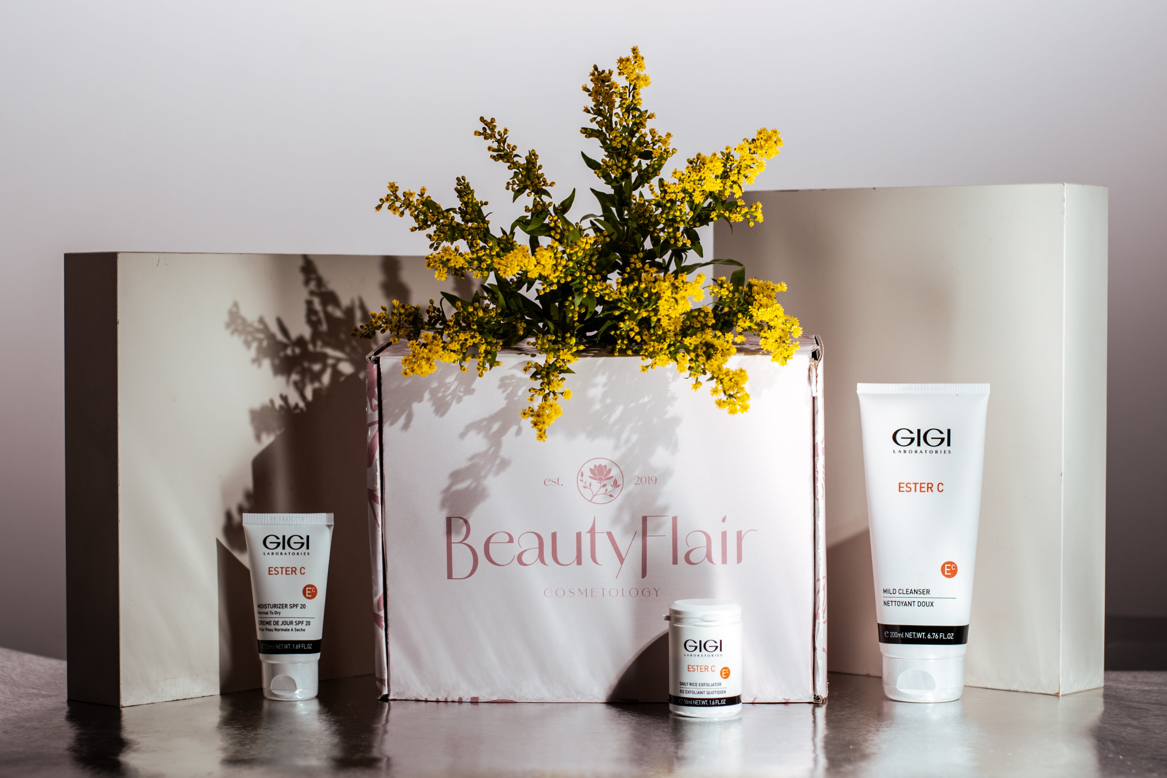 BeautyFlair kosmeetikakarp kollaste lilledega, milles on GIGI ESTER C sarja tooted: niisutav kreem SPF 20, näopuhastusvahend ja kreemipurk.